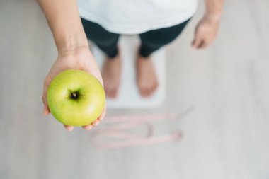 Ağırlık ölçeğinde bir kadın ve el ele tutuşan yeşil elma, meyve seçmek sağlıklı yemektir. Diyet kontrolü, kilo kaybı, obezite, yeme tarzı ve beslenme kavramları