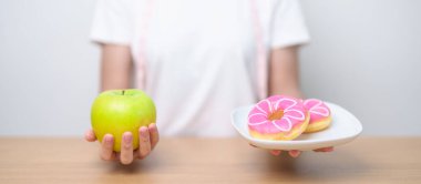 Kadın elini yeşil elma ve çörek ile tutar, kadın fitness meyve ve tatlı sağlıklı ve sağlıksız abur cubur arasında seçim yapar. Diyet kontrolü, kilo kaybı, obezite, yeme tarzı ve beslenme konsepti