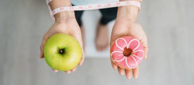Ağırlık ölçeğinde ve elinde yeşil elma ve çörek olan bir kadın, meyve ve tatlı arasında seçim yapmak sağlıksız ve abur cuburdur. Diyet kontrolü, kilo kaybı, obezite, yeme tarzı ve beslenme