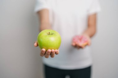 Kadın elini yeşil elma ve çörek ile tutar, kadın fitness meyve ve tatlı sağlıklı ve sağlıksız abur cubur arasında seçim yapar. Diyet kontrolü, kilo kaybı, obezite, yeme tarzı ve beslenme konsepti