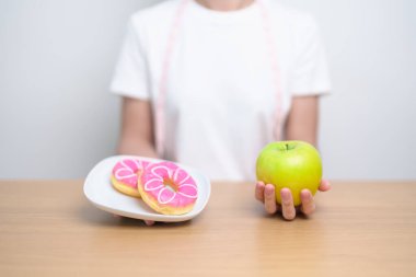 Kadın elini yeşil elma ve çörek ile tutar, kadın fitness meyve ve tatlı sağlıklı ve sağlıksız abur cubur arasında seçim yapar. Diyet kontrolü, kilo kaybı, obezite, yeme tarzı ve beslenme konsepti