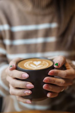 Kışın kalp latte sanatıyla kapuçino içen genç bir kadın, Noel tırnak sanatı manikürlü bir kadın ve sabahları evde bir fincan kahvenin keyfini çıkarıyor.