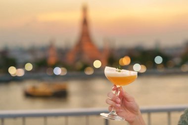 tourist hand shows cocktail glass during enjoy view to Wat Arun Temple in sunset, Traveler visits Temple of Dawn near from rooftop bar. Travel destination in Bangkok, Thailand and Southeast Asia clipart