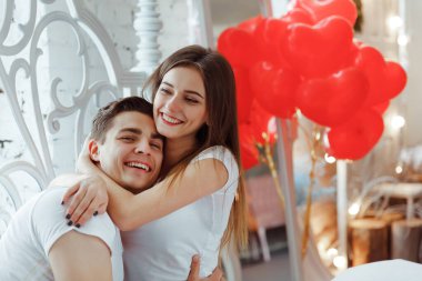 Evdeki güzel genç çift. Sevgililer Günü 'nü arka planda kalp şeklinde balonlarla kutlarken sarılıp öpüşmek ve birlikte vakit geçirmekten zevk almak..