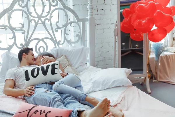 Beautiful young couple in bedroom is lying on bed with air balloons in shape of heart. Enjoying spending time together. Celebrating Saint Valentines Day.