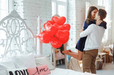 Güzel genç çift evde. Sarılma, öpüşme ve birlikte üzerinde kalp şeklinde yatak ve hava balonları ve arka plan üzerinde kırmızı gül ile Saint Valentine's Day kutlamaları sırasında vakit zevk.