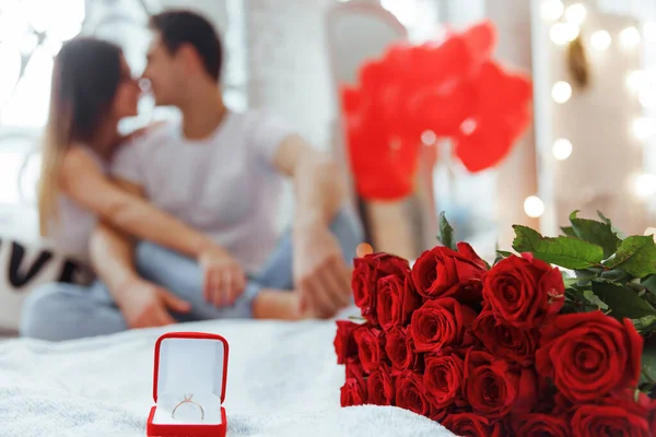 Proposal ring and red roses on the bed with couple in love on background.Man makes present to his lovely sweetheart girl. Valentine Couple. Family. Boy gives to his girlfriend jewelry.