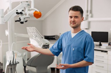 Beyaz önlüklü yakışıklı dişçi kameraya bakıyor ve ofisini gösteriyor.