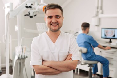 Arka planda çalışan asistanıyla dişçi kliniğinde kollarını kavuşturmuş gülümseyen bir dişçinin portresi.