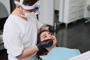 Diş hekimi ağız aynası ve diş kazımıyla hasta dişlerini inceliyor. Kadının yüzüne yakından bak.