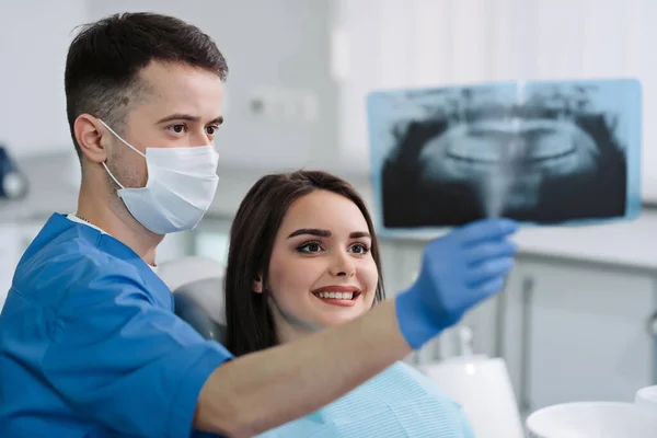 Dentist with mask in uniform and beautiful female patient looking at dental x-ray.Modern dental office. Dental treatment.