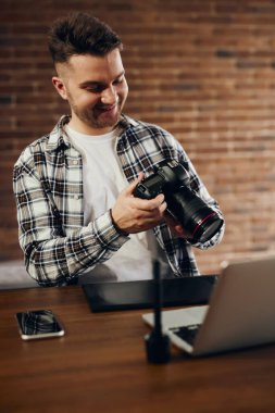 A professional photographer holds a camera in his hands and sits at his office desk. There is a laptop and a graphic tablet on the table.