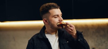 Handsome young man enjoying pizza in his kitchen. Unbelievably enjoys the taste.