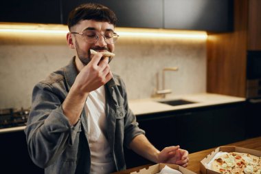 A handsome young man in glasses is enjoying pizza in his kitchen. Unbelievably enjoys the taste.