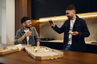 Two guys in the kitchen talking and eating pizza and drinking beer. Male friendship.