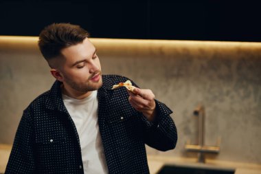 Handsome young man enjoying pizza in his kitchen. Unbelievably enjoys the taste.