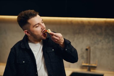 Handsome young man enjoying pizza in his kitchen. Unbelievably enjoys the taste.
