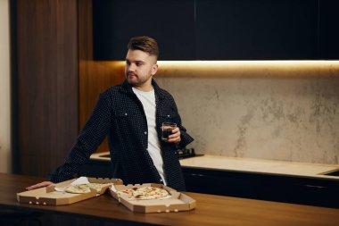 Handsome young man enjoying pizza in his kitchen and drinking beer.