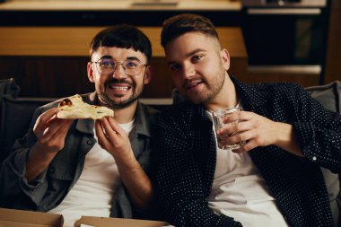 Two happy young guys are eating pizza and watching TV together. The concept of friendly outings watching TV series, sports competition, football.
