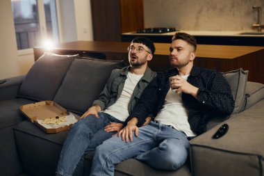 Two men are watching TV and eating pizza. Emotion of intrigue, interest.