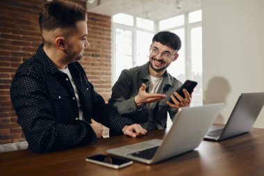 Two happy young men are working in the office on laptops. One shows the other something on the phone.