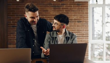 Two happy young men are working in the office on laptops. One of them shows the other how to do something correctly.