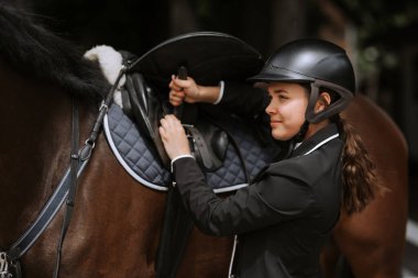 Kız binici at yarışlarına katılmak için atının eyer ayarlar.