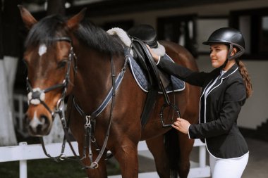 Kız binici at yarışlarına katılmak için atının eyer ayarlar.