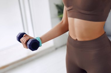 A model is warming up with dumbbell while standing on the mat. She has a light-blue clock and dark dumbbell. She is wearing clothes for fitness.