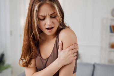 A yoga instructor stretches her left arm with her right hand, her facial expression painful. She is looking at her left hand. There is a gray sofa near her. She is also wearing brown clothes. She has a light-brown hair.
