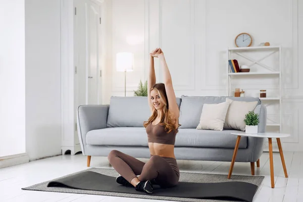 Fitness woman exercising and stretching arms at home in the living room in sportswear. Female doing relaxing yoga and breathing exercises.