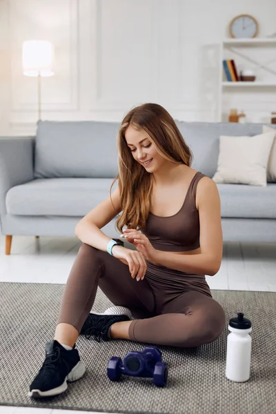 Young beautiful athletic girl in leggings and top does stretching exercises. Healthy lifestyle. A woman using smart watch and goes in for sports at home.