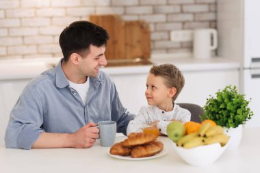 Father and son are talking and smiling while having a breakfast on kitchen.