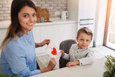 Young woman with her little son and greeting card for Mother's Day at home. Happy woman receiving flowers and greeting card from her son at home. Mother's day celebration