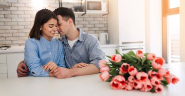 Happy beautiful cute young couple are sitting together on the kitchen with flowers. Love, celebration, romantic, st. Valentine's day, women day, birthday, date.