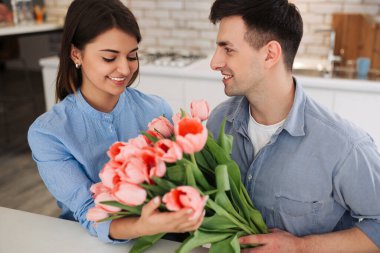 Surprise! Beautiful romantic couple at home. Young man is presenting flowers to his beloved. Feel of happiness