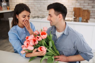 Surprise! Beautiful romantic couple at home. Young man is presenting flowers to his beloved. Feel of happiness.