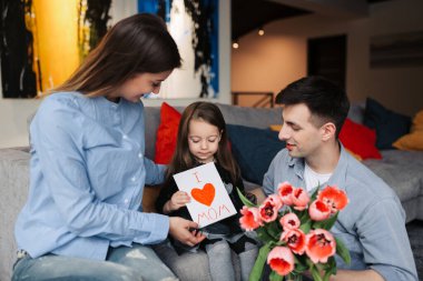 Happy mother's day! Child daughter and father is congratulating mom and giving her postcard and gift. Family smiling and hugging. Family holiday and togetherness