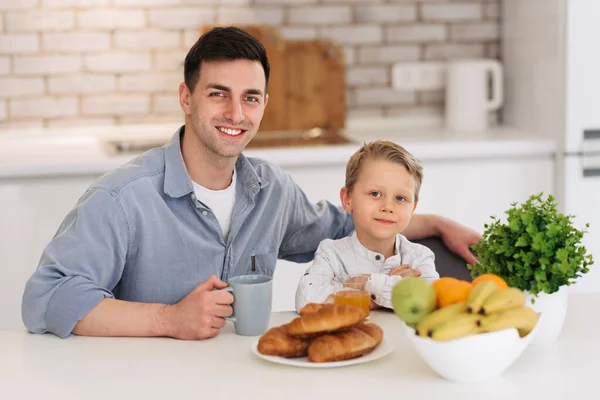 Father and son talking and smiling while having breakfast in the kitchen. They look into the camera and smile. Happy family concept