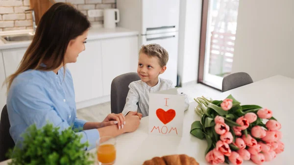 Cute little boy greeting his mother at home. Mother\'s day concept. Happy mother\'s day! Child son congratulates moms and gives her a postcard and flowers