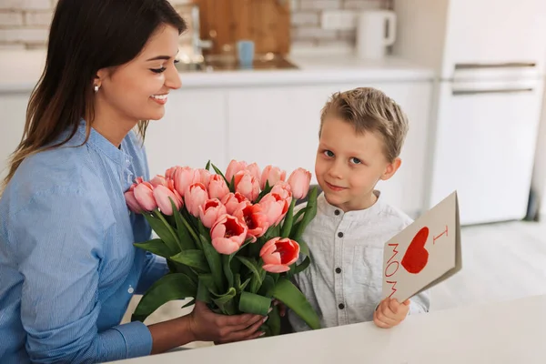 Young woman with her little son and greeting card for Mother\'s Day at home. Happy woman receiving flowers and greeting card from her son at home. Mother\'s day celebration