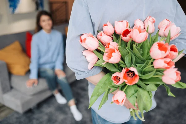 Unexpected moment in routine everyday life! Cropped photo of man\'s hands hiding holding chic bouquet of tulips behind back, happy woman is on background.