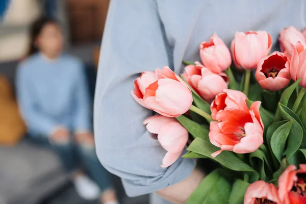 Unexpected moment in routine everyday life! Cropped photo of man\'s hands hiding holding chic bouquet of tulips behind back, happy woman is on background