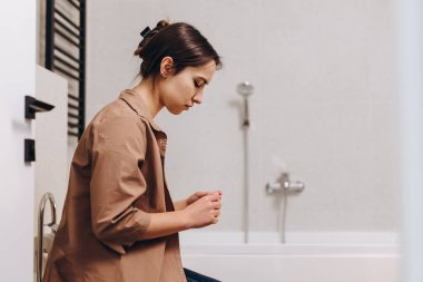 A young worried girl is sitting in the bathroom and holding a pregnancy test in her hands. She is waiting and afraid of the unknown.