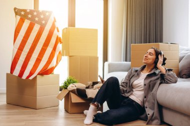 A young teenager has moved to a new apartment and is listening to music in headphones. The concept of happy moving, studentship. The USA flag is in the background.