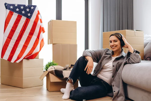 A young teenager has moved to a new apartment and is listening to music in headphones. The concept of happy moving, studentship. The USA flag is in the background.