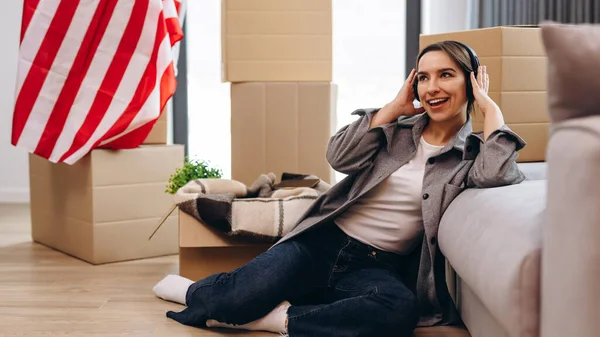 A young teenager has moved to a new apartment and is listening to music in headphones. The concept of happy moving, studentship. The USA flag is in the background.