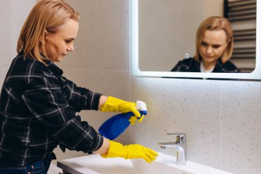 An elderly woman washes the sink in the bathroom.