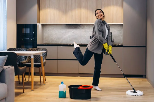 Funny teenager is cleaning house doing housework cleaning floor with mop and listening to music in wireless headphones, girl is dancing and singing.