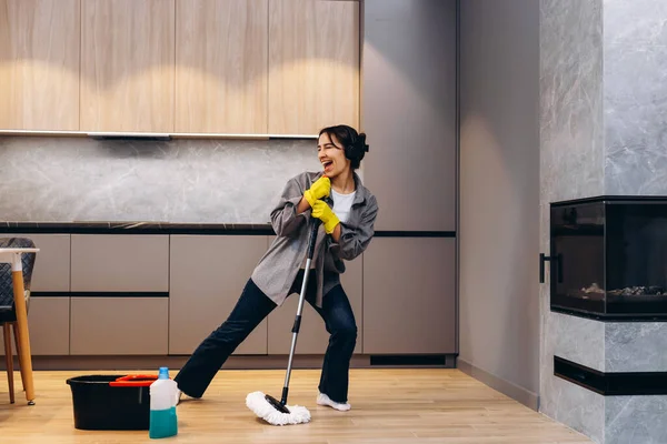 Funny teenager is cleaning house doing housework cleaning floor with mop and listening to music in wireless headphones, girl is dancing and singing.